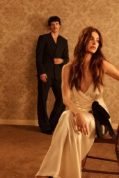 Camila Morrone posing in cream silk dress with black bow and black leather dress in Netflix photoshoot with ornate damask wallpaper backdrop, April 2026