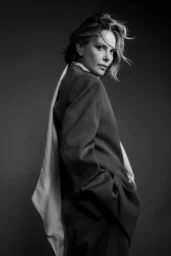 Charlize Theron posing for New York Times photoshoot April 2026, wearing dark blazer with white shirt and transparent raincoat with belt, wind-blown hair, black and white studio portrait aesthetic