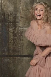 Claudia Black posing in white embellished gown, gold tweed jacket with cream trousers, and pink ruffled dress for Glamour BG Magazine "Beyond the Stars" editorial photoshoot, March 2026, romantic styling with blonde curly haira