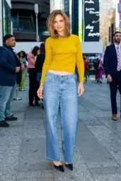 Katie Holmes standing in Times Square April 2026 wearing a cropped yellow sweater with light blue wide leg jeans and black pointed shoes