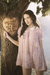 Olivia Rodrigo posing in pink floral dress next to tree with carved heart for 2026 photoshoot, romantic outdoor setting with natural lighting